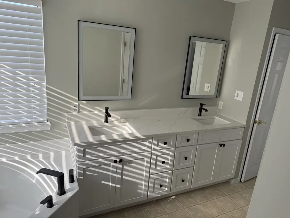 White double vanity with matte black faucets and two framed mirrors, morning light through the blinds