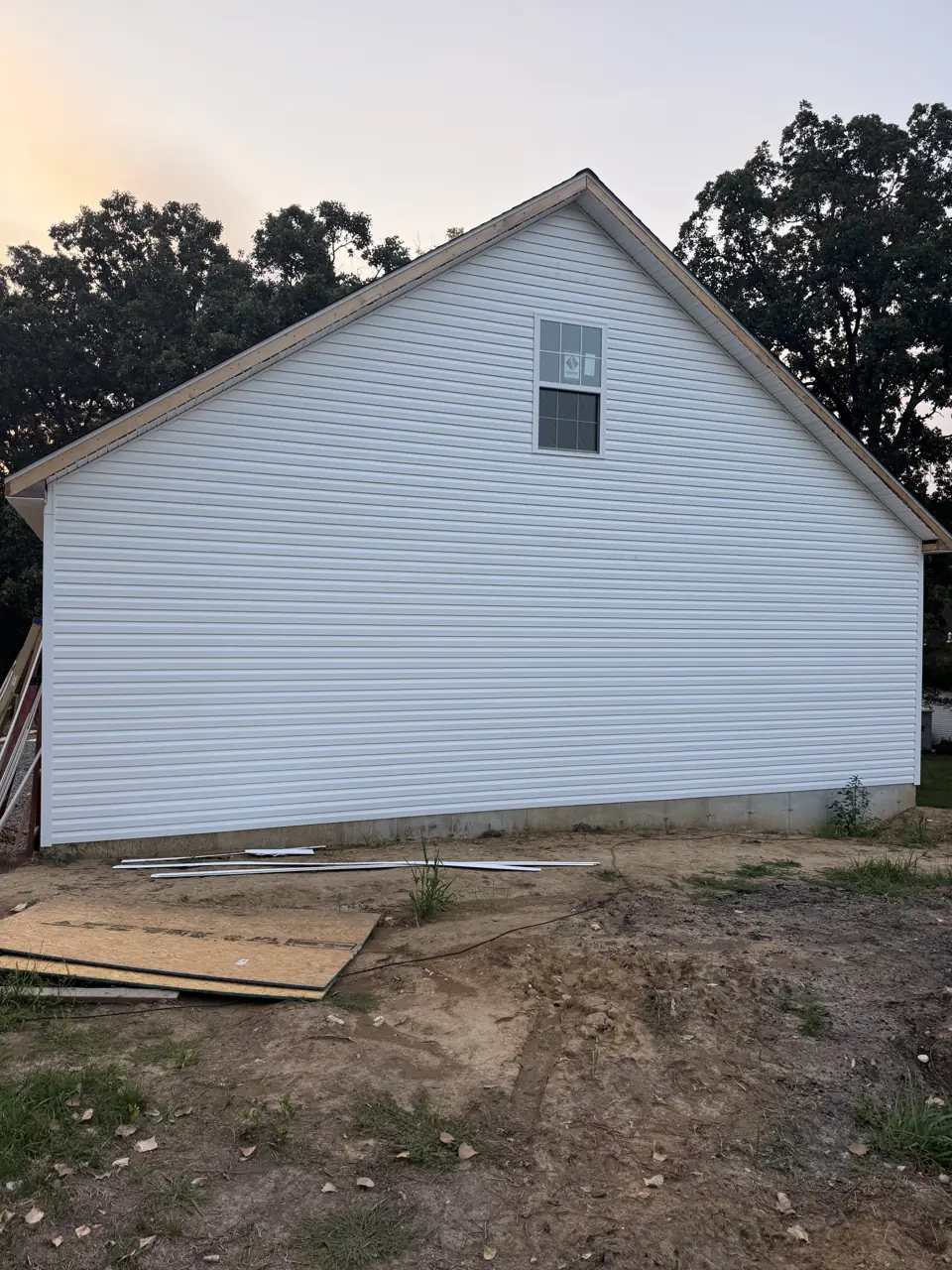 Finished gable end of a new detached garage with white siding and a small window, trees behind at dusk