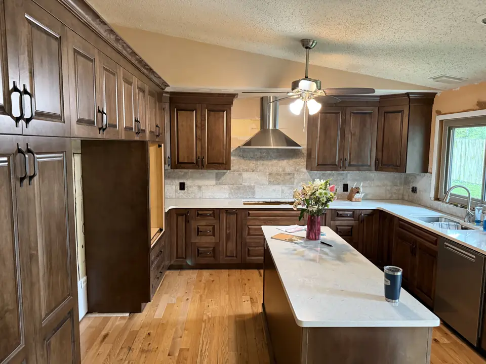 Tall stained pantry cabinets beside a kitchen with white quartz island and travertine backsplash