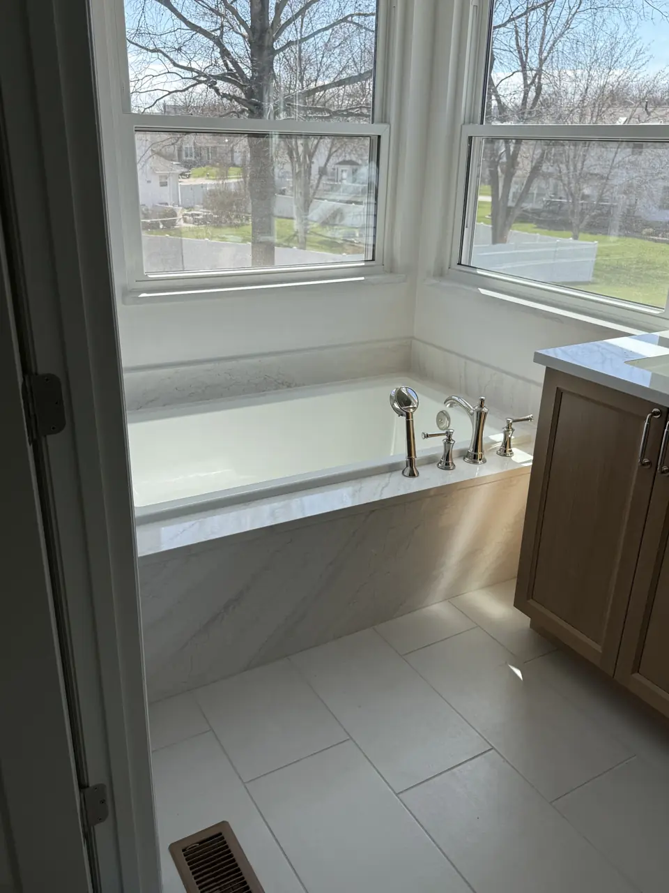 Drop-in soaking tub set under a bay window with a porcelain slab surround and a light oak vanity