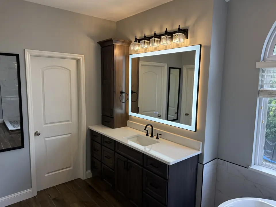 Dark vanity with white counter and a large backlit LED mirror in a grey bathroom with wood-look plank tile