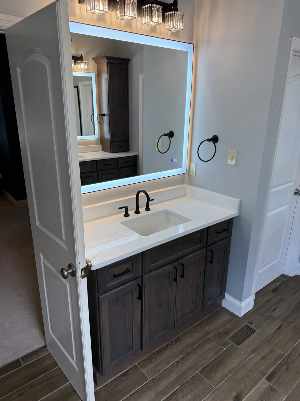 Backlit LED vanity mirror over a dark single vanity with matte black faucet, seen through the bathroom doorway