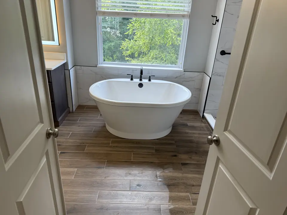 White freestanding soaking tub in a window alcove with a matte black floor-mounted filler and wood-look tile floor