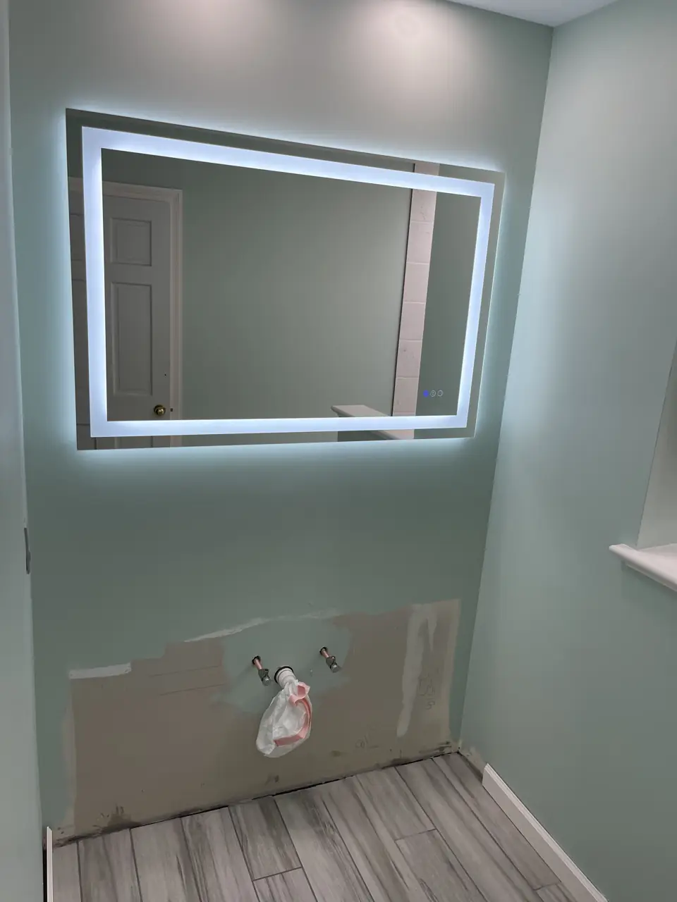 Backlit LED mirror glowing on a sage green wall beside the bathroom window