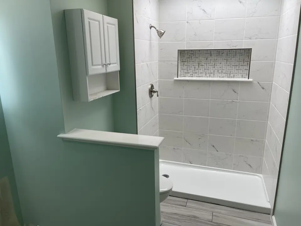 Tiled shower with a framed niche and knee wall beside a white medicine cabinet on a sage green wall