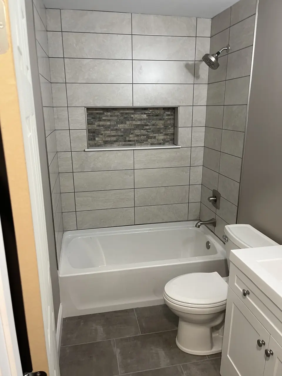 Bathtub with large-format grey tile surround in a staggered pattern, a glass mosaic niche, dark floor tile and a white vanity