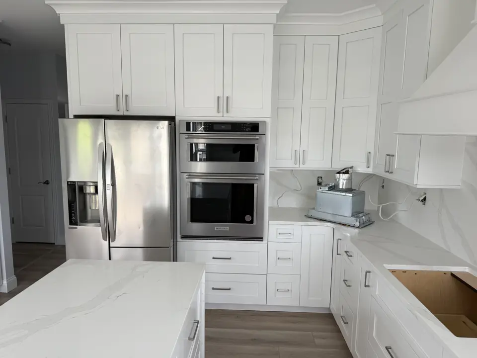 White shaker kitchen with stainless refrigerator and wall ovens set into a full-height cabinet run beside a quartz island